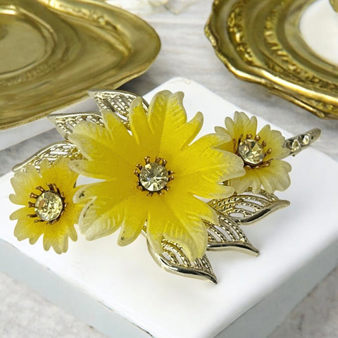 Vintage yellow celluloid floral brooch from the 1950s featuring three gradient yellow-to-white flowers with clear rhinestone centers, accented by light gold-tone openwork leaves.