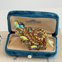 Gold‑tone vintage brooch with AB topaz, smoky, and clear rhinestones in a floral swirl shape.