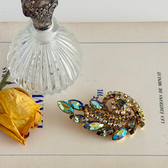 Gold‑tone vintage brooch with AB topaz, smoky, and clear rhinestones in a floral swirl shape.