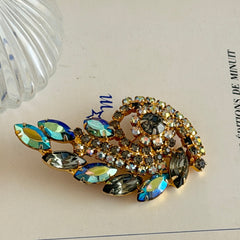Gold‑tone vintage brooch with AB topaz, smoky, and clear rhinestones in a floral swirl shape.
