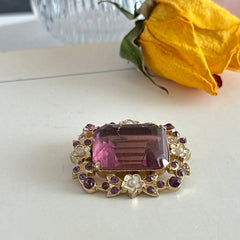 Vintage gold‑tone brooch with an amethyst‑colored rhinestone, faux pearls, and white enamel flowers