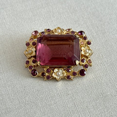 Vintage gold‑tone brooch with an amethyst‑colored rhinestone, faux pearls, and white enamel flowers