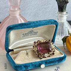 Vintage gold‑tone brooch with an amethyst‑colored rhinestone, faux pearls, and white enamel flowers