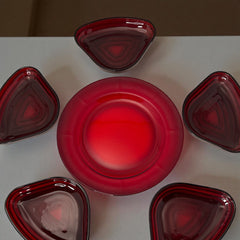 Vintage Anchor Hocking Royal Ruby relish trays arranged in a circular pattern around a coordinating red glass plate. The translucent ruby red glass glows with depth and symmetry, showcasing the sculptural triangular shapes and rich mid-century color. Ideal for holiday styling or vintage table settings.
