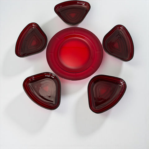 Vintage Anchor Hocking Royal Ruby relish trays arranged in a circular pattern around a coordinating red glass plate. The translucent ruby red glass glows with depth and symmetry, showcasing the sculptural triangular shapes and rich mid-century color. Ideal for holiday styling or vintage table settings.