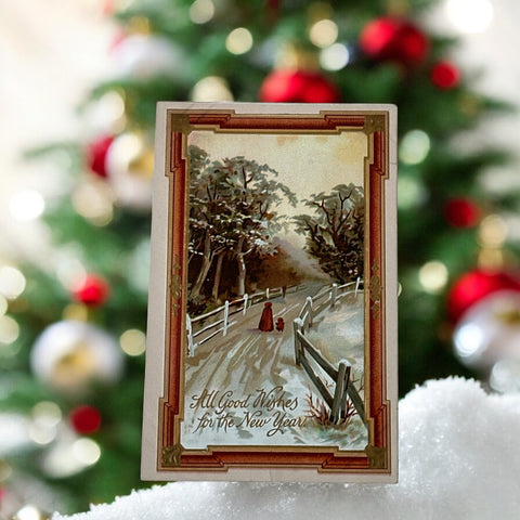 Antique 1910s embossed postcard featuring a snowy path with a person and child walking, framed by gilded orange-brown border; message reads “All Good Wishes for the New Year.