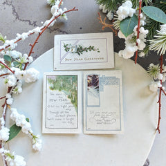 Three vintage New Year postcards from the 1920s arranged on a wooden surface: one embossed with holly and a gold calendar motif, one printed with a poetic candle verse, and one featuring a winter forest scene with ornate blue border.