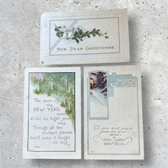 Three vintage New Year postcards from the 1920s arranged on a wooden surface: one embossed with holly and a gold calendar motif, one printed with a poetic candle verse, and one featuring a winter forest scene with ornate blue border.