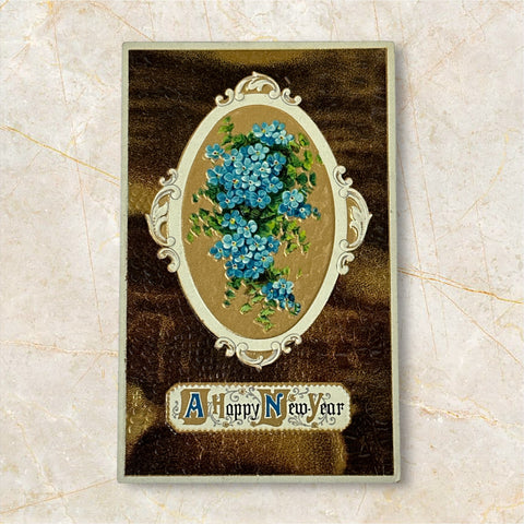 Antique 1910s New Year postcard featuring embossed blue forget-me-not flowers in a gold-gilded oval frame, set against a textured brown background resembling crocodile skin; elegant and well-preserved with handwritten 1913 message on reverse.