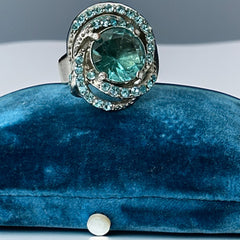 Adjustable silver-tone cocktail ring with a large round aqua rhinestone cabochon and open swirl design set with smaller teal rhinestones; vintage-inspired statement jewelry in very good condition.