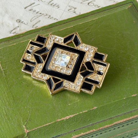 Vintage Art Deco style gold‑tone brooch with black enamel and clear rhinestones, geometric design photographed on antique French script background.