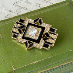 Vintage Art Deco style gold‑tone brooch with black enamel and clear rhinestones, geometric design photographed on antique French script background.