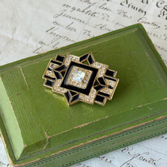 Vintage Art Deco style gold‑tone brooch with black enamel and clear rhinestones, geometric design photographed on antique French script background.