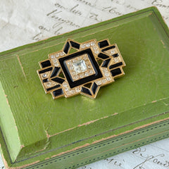 Vintage Art Deco style gold‑tone brooch with black enamel and clear rhinestones, geometric design photographed on antique French script background.