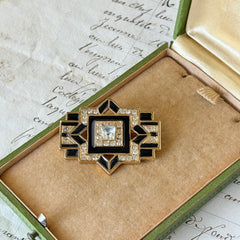 Vintage Art Deco style gold‑tone brooch with black enamel and clear rhinestones, geometric design photographed on antique French script background.