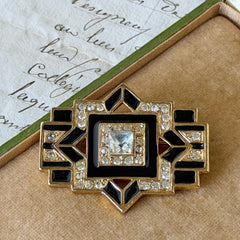 Vintage Art Deco style gold‑tone brooch with black enamel and clear rhinestones, geometric design photographed on antique French script background.