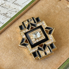 Vintage Art Deco style gold‑tone brooch with black enamel and clear rhinestones, geometric design photographed on antique French script background.