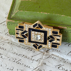 Vintage Art Deco style gold‑tone brooch with black enamel and clear rhinestones, geometric design photographed on antique French script background.