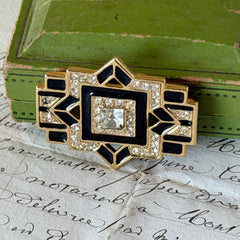 Vintage Art Deco style gold‑tone brooch with black enamel and clear rhinestones, geometric design photographed on antique French script background.