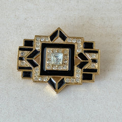 Vintage Art Deco style gold‑tone brooch with black enamel and clear rhinestones, geometric design photographed on antique French script background.