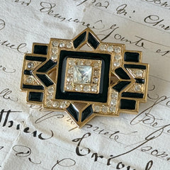 Vintage Art Deco style gold‑tone brooch with black enamel and clear rhinestones, geometric design photographed on antique French script background.