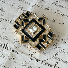Vintage Art Deco style gold‑tone brooch with black enamel and clear rhinestones, geometric design photographed on antique French script background.