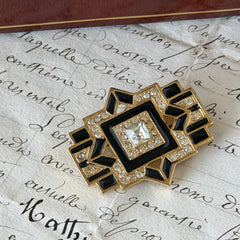 Vintage Art Deco style gold‑tone brooch with black enamel and clear rhinestones, geometric design photographed on antique French script background.