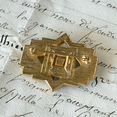 Vintage Art Deco style gold‑tone brooch with black enamel and clear rhinestones, geometric design photographed on antique French script background.