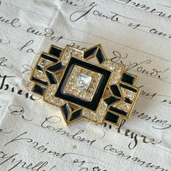 Vintage Art Deco style gold‑tone brooch with black enamel and clear rhinestones, geometric design photographed on antique French script background.