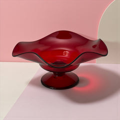 Vintage ruby red hand-blown glass bowl with a freeform wavy rim and clear pedestal base, photographed from above to highlight its organic curves and glossy surface. Midcentury compote style, ideal for decorative or serving use.