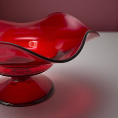 Vintage ruby red hand-blown glass bowl with a freeform wavy rim and clear pedestal base, photographed from above to highlight its organic curves and glossy surface. Midcentury compote style, ideal for decorative or serving use.