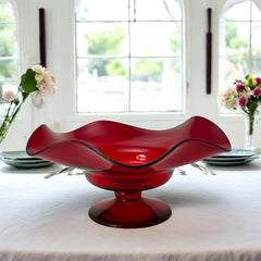Vintage ruby red hand-blown glass bowl with a freeform wavy rim and clear pedestal base, photographed from above to highlight its organic curves and glossy surface. Midcentury compote style, ideal for decorative or serving use.