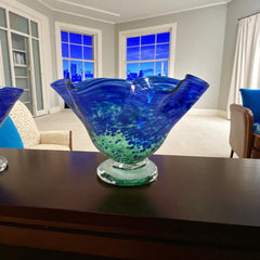 Signed BAGI art glass bowl with a footed base and wavy rim, featuring mottled blue and green swirls. The glass has a glossy finish and sculptural form, with vibrant oceanic and forest tones. Dimensions approximately 5–5½" tall and 7–8" wide.