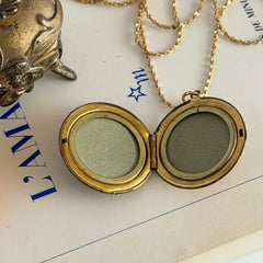 Round vintage black enamel locket with raised gold floral motif and engraved gold‑tone border on a delicate tinsel chain.