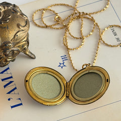 Round vintage black enamel locket with raised gold floral motif and engraved gold‑tone border on a delicate tinsel chain.
