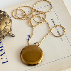 Round vintage black enamel locket with raised gold floral motif and engraved gold‑tone border on a delicate tinsel chain.