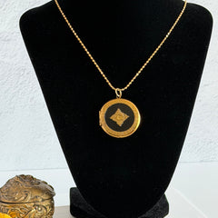 Round vintage black enamel locket with raised gold floral motif and engraved gold‑tone border on a delicate tinsel chain.