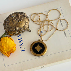 Round vintage black enamel locket with raised gold floral motif and engraved gold‑tone border on a delicate tinsel chain.