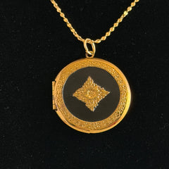 Round vintage black enamel locket with raised gold floral motif and engraved gold‑tone border on a delicate tinsel chain.