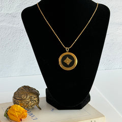 Round vintage black enamel locket with raised gold floral motif and engraved gold‑tone border on a delicate tinsel chain.