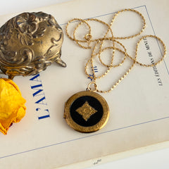 Round vintage black enamel locket with raised gold floral motif and engraved gold‑tone border on a delicate tinsel chain.