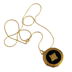 Round vintage black enamel locket with raised gold floral motif and engraved gold‑tone border on a delicate tinsel chain.