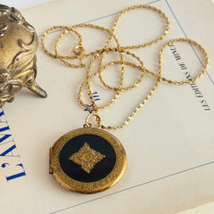 Round vintage black enamel locket with raised gold floral motif and engraved gold‑tone border on a delicate tinsel chain.