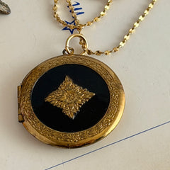 Round vintage black enamel locket with raised gold floral motif and engraved gold‑tone border on a delicate tinsel chain.