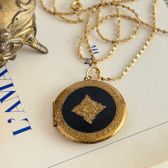 Round vintage black enamel locket with raised gold floral motif and engraved gold‑tone border on a delicate tinsel chain.