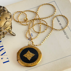 Round vintage black enamel locket with raised gold floral motif and engraved gold‑tone border on a delicate tinsel chain.