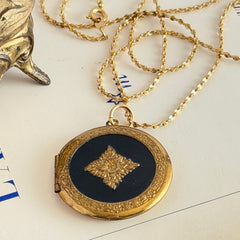 Round vintage black enamel locket with raised gold floral motif and engraved gold‑tone border on a delicate tinsel chain.