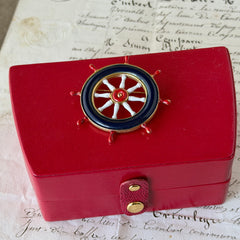 Vintage gold‑tone ship‑wheel brooch with navy, red, and white enamel, photographed on antique French script background.