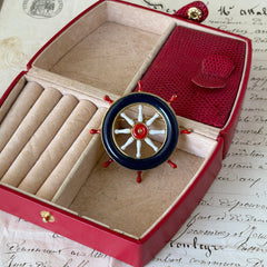 Vintage gold‑tone ship‑wheel brooch with navy, red, and white enamel, photographed on antique French script background.
