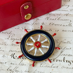 Vintage gold‑tone ship‑wheel brooch with navy, red, and white enamel, photographed on antique French script background.